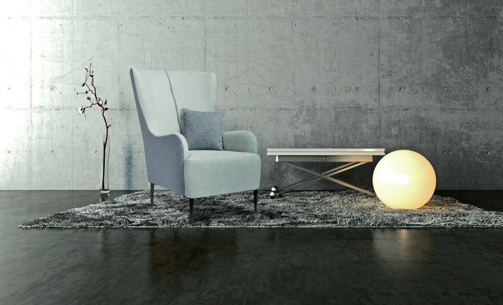 Fauteuil à oreilles Harry en tissu gris clair avec coussin, pieds en métal noir, devant un mur de béton